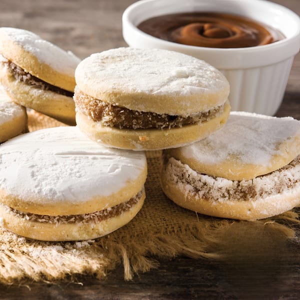 Peruvian alfajores, powdered sugar cookies with dulce de leche filling, on a burlap cloth with a bowl of caramel.
