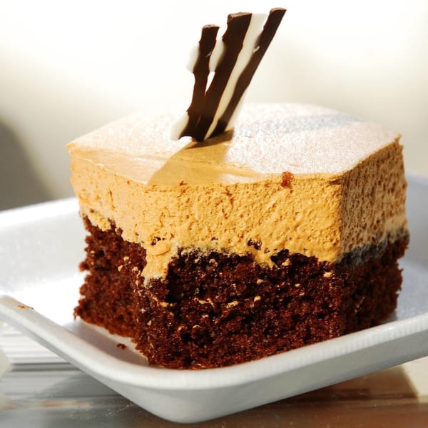A square slice of chocolate cake with a thick layer of light brown mousse, topped with chocolate shards.